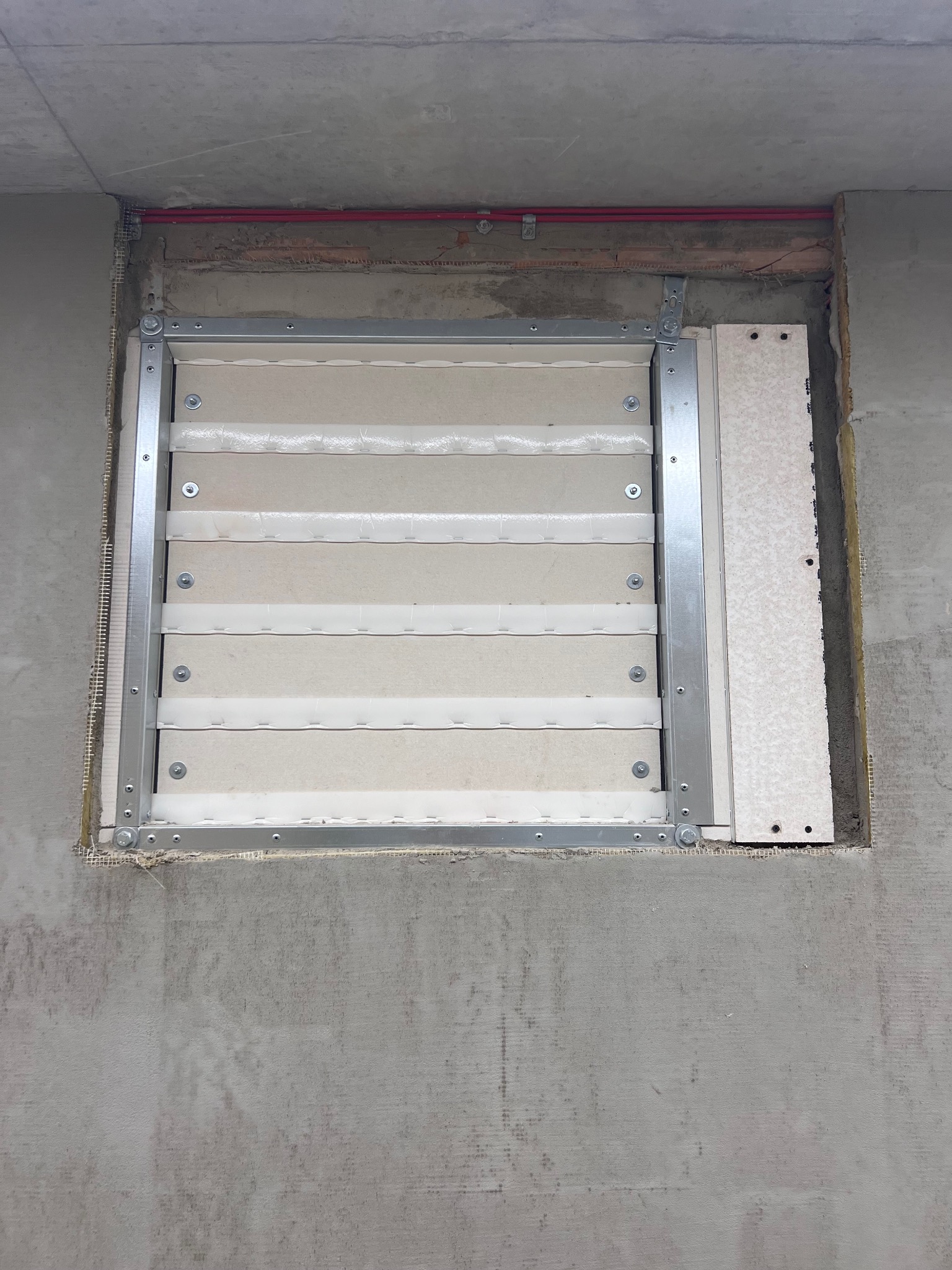 Zamontowana kratka wentylacyjna z metalową ramą i poziomymi lamelami, widoczna w surowym otworze w ścianie z betonu komórkowego, z widoczną izolacją i siatką zbrojeniową.