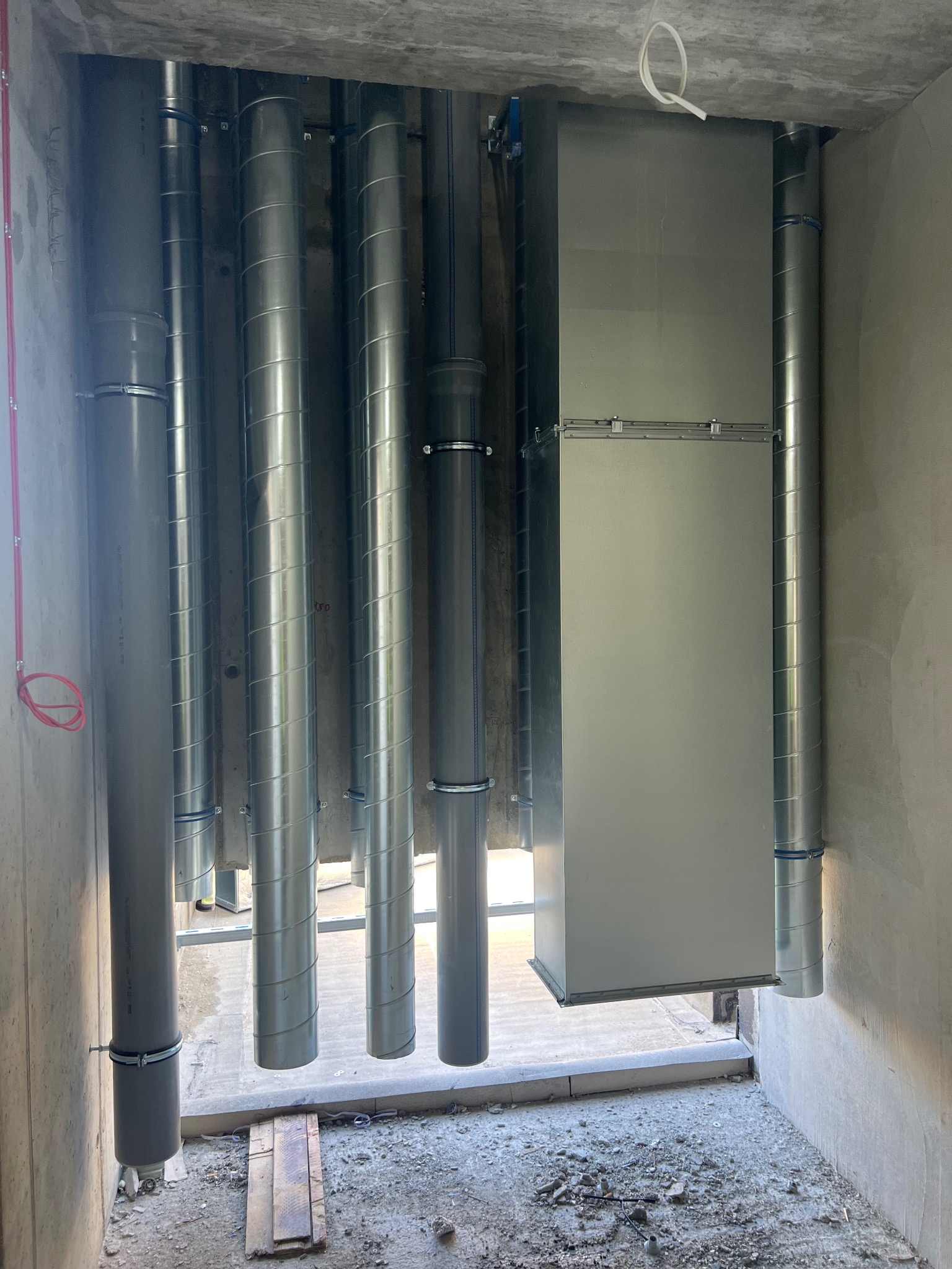 Wnętrze w trakcie instalacji systemu wentylacyjnego z widocznymi rurami spiralnymi, prostokątnym kanałem wentylacyjnym i rurami PCV, zamocowanymi do surowego betonowego stropu.