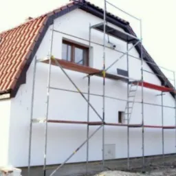 Dom jednorodzinny w trakcie ocieplania, widoczne rusztowania i świeżo otynkowana biała elewacja, czerwona dachówka.