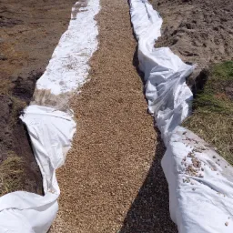 Przygotowanie podłoża pod nawierzchnię z drobnego kruszywa, z użyciem białej geowłókniny w wykopie ziemnym; widoczni pracownicy w oddali.