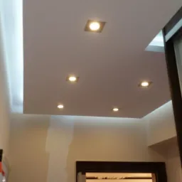 Podwieszany sufit z oświetleniem punktowym w jasnym wnętrzu. Widoczne fragmenty ścian i drzwi. Minimalistyczny design.