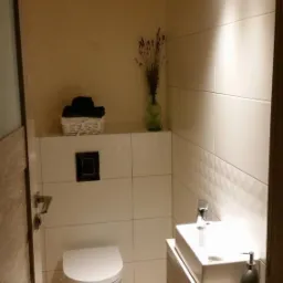 Nowoczesna łazienka z białymi płytkami, podwieszaną toaletą i umywalką na szafce. Wazon z suszem i koszyk z ręcznikami dodają przytulności.