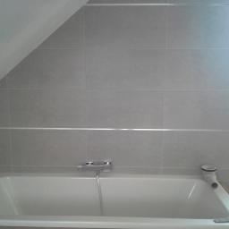 Jacek Rzepka Usługi Remontowo Budowlane - Wykończona ściana nad wanną w łazience na poddaszu, wyłożona szarymi, kwadratowymi płytkami z aluminiową listwą dekoracyjną. Widoczna bateria i odpływ wanny.