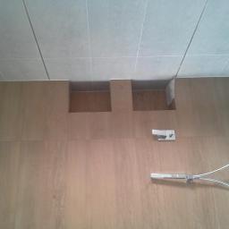 Jacek Rzepka Usługi Remontowo Budowlane - Wykończona ściana prysznicowa z drewnopodobnych płytek, wnękami i armaturą. Widok z dołu, lekko obrócony. Jasne płytki na suficie.