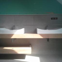 Jacek Rzepka Usługi Remontowo Budowlane - Wykończona łazienka z dwiema białymi umywalkami nablatowymi na drewnianym blacie. Szare płytki ścienne i podłogowe. Widoczne gniazdka elektryczne i płyta karton-gips.