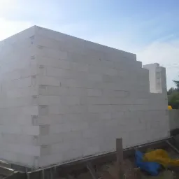 Wznoszenie ścian domu z bloczków betonu komórkowego Ytong, widoczne fragmenty fundamentów i zarys przyszłych pomieszczeń, błękitne niebo w tle.