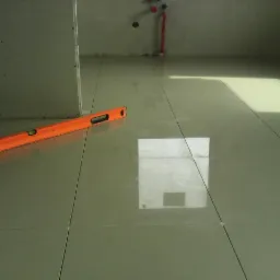 Połyskujące, beżowe płytki podłogowe w trakcie układania, widoczna poziomica i fragment ściany z płyt karton-gips w łazience. Oświetlenie naturalne.