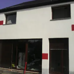 Biała fasada budynku z czerwonymi akcentami. Widoczne okna z brązowymi ramami i częściowo zasłonięte rolety. Na lewo rusztowanie.