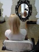 Klientka siedzi na fotelu w salonie, widok z tyłu na blond włosy przed zabiegiem przedłużania, w tle lustro w ozdobnej ramie.