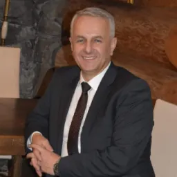 Uśmiechnięty mężczyzna w garniturze siedzi przy drewnianym stole, w tle kamienna ściana, lampa i obraz z motywem wiejskim.