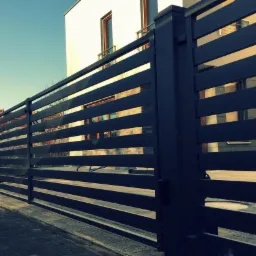 Nowoczesne, poziome ogrodzenie panelowe w kolorze grafitowym, z widocznymi budynkami w tle i kostką brukową na pierwszym planie. Ujęcie z niskiej perspektywy.