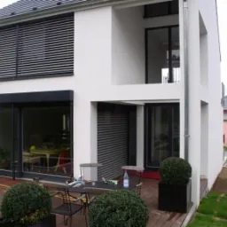 Nowoczesny dom z białymi ścianami, dużymi przesuwnymi oknami z roletami zewnętrznymi, widok z tarasu na drewnianym podeście, na którym stoi stół z krzesłami i narzędziami.