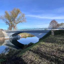 Nowy betonowy most nad spokojną rzeką w słoneczny dzień, odbicie konstrukcji w wodzie, drzewa w tle, świeżo obsiana trawa na brzegu.