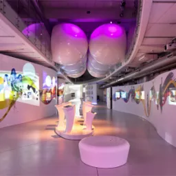 Nowoczesna przestrzeń wystawiennicza z interaktywnymi ekranami na zakrzywionych ścianach, oświetlona fioletowym światłem, z futurystycznymi meblami i podwieszanymi elementami dekoracyjnymi.