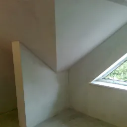 Poddasze po tynkowaniu maszynowym, widoczne ściany i skośny sufit z oknem trójkątnym, nierówności na powierzchni przed malowaniem.