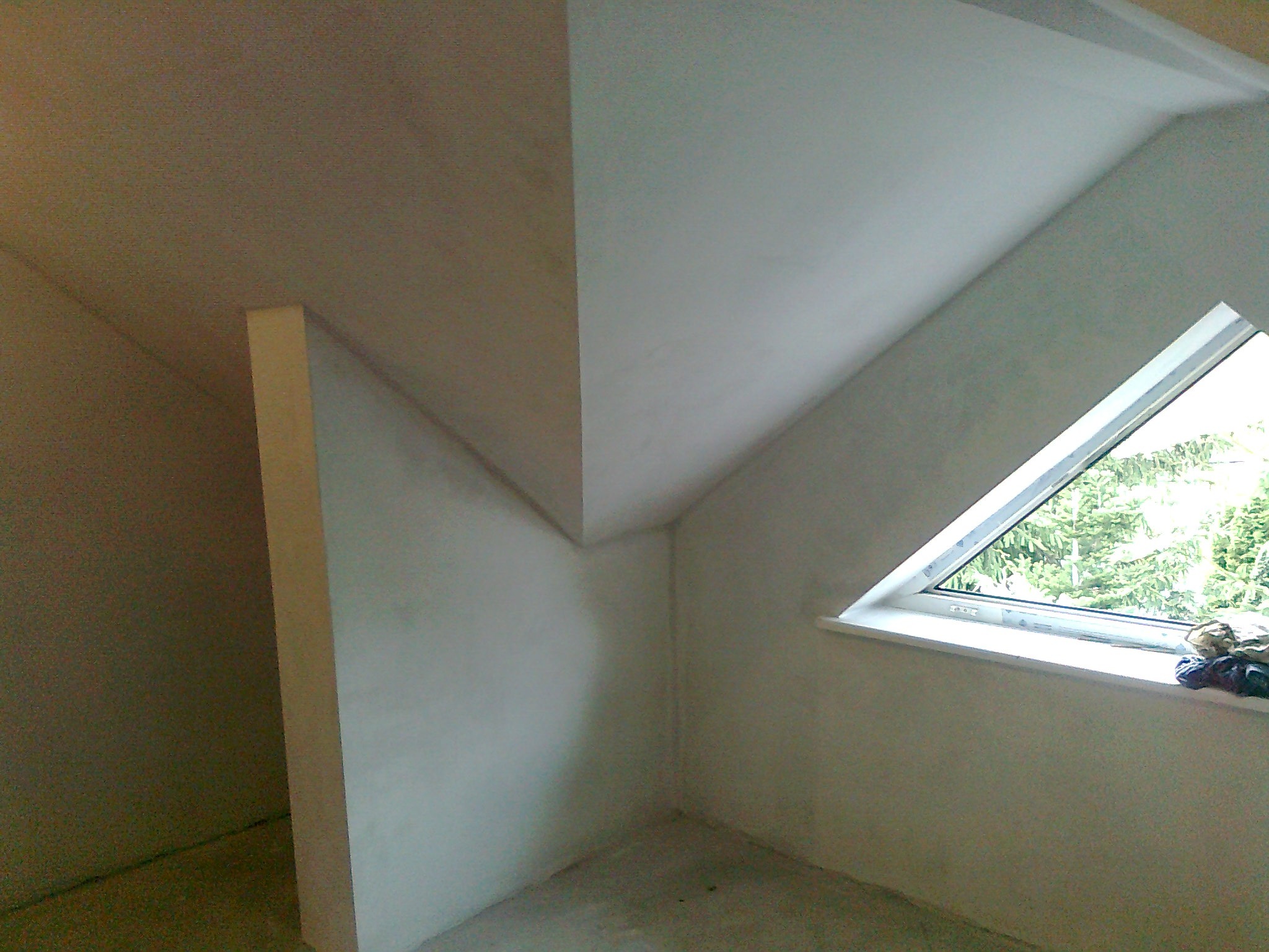 Poddasze po tynkowaniu maszynowym, widoczne ściany i skośny sufit z oknem trójkątnym, nierówności na powierzchni przed malowaniem.