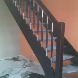 Czarne schody z metalowymi balustradami w trakcie montażu, narzędzia i elementy leżą na podłodze zabezpieczonej folią. Pomarańczowa ściana w tle.