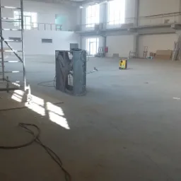 Wnętrze hali w trakcie budowy lub remontu, widoczny duży wentylator przemysłowy, agregat prądotwórczy oraz rusztowanie. Podłoga betonowa, surowe ściany i strop.