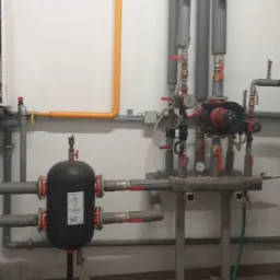 Wnętrze kotłowni z otwartym kotłem gazowym, zbiornikiem wyrównawczym, pompami obiegowymi i szarą instalacją rurową z zaworami, widoczne elementy drabiny malarskiej.