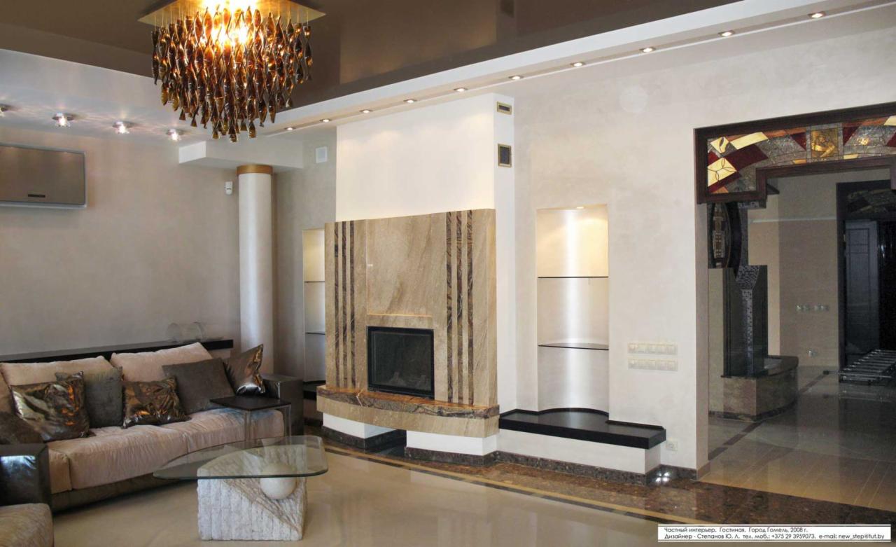 Elegancki salon z marmurowym kominkiem i designerskim żyrandolem. Beżowe ściany, sofa z poduszkami i szklany stolik kawowy tworzą luksusową przestrzeń. Widoczne detale wykończenia.