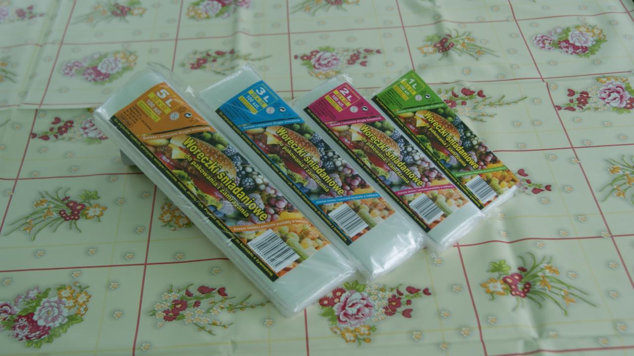 Zestaw czterech rolek woreczków śniadaniowych różnych pojemności (1L, 2L, 3L, 5L) ułożonych na kwiecistym obrusie, przeznaczonych do pakowania i zamrażania żywności.