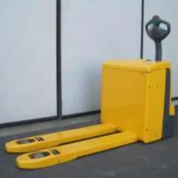 Żółty, używany wózek paletowy prowadzony dyszlem, z widocznymi wyświetlaczami na widłach, stojący na szarym podłożu przed jasnymi drzwiami garażowymi.