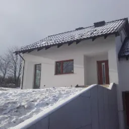 Biały dom jednorodzinny z brązowymi oknami i dachem pokrytym śniegiem, widziany z dołu w zimowej scenerii.