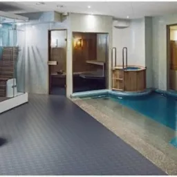 Luksusowe wnętrze spa z basenem, jacuzzi w drewnianej obudowie i szklaną kabiną prysznicową z drewnianym siedziskiem. Błękitna mozaika na ścianach i szara podłoga.