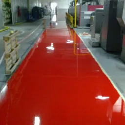 Błyszcząca, czerwona posadzka przemysłowa w hali produkcyjnej, świeżo po nałożeniu. Widoczne palety i maszyny w tle, podkreślające przemysłowy charakter.