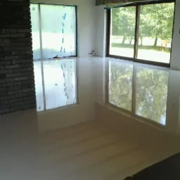 Błyszcząca, jasna posadzka po sprzątaniu po remoncie, z widoczną ceglaną ścianą i zabezpieczonymi oknami folią ochronną. Duże okna z widokiem na zieleń.