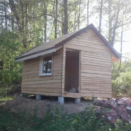 Surowy drewniany domek letniskowy w Koszalinie, z widocznym pojedynczym oknem i stertą dachówek obok, otoczony zielenią.