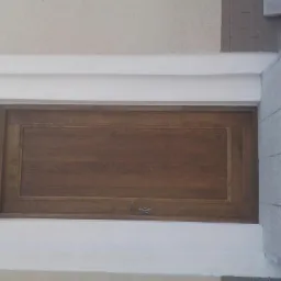 Zamknięte, drewniane drzwi garażowe z poziomym usłojeniem, osadzone w białej ramie, z widocznym fragmentem szarej, betonowej ściany obok.