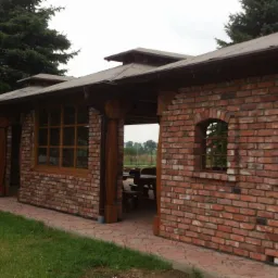 Zadaszona konstrukcja z cegły z drewnianymi oknami i filarami, widok z zewnątrz na trawnik i drzewa.