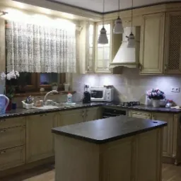 Klasyczna kuchnia z wyspą, kremowe szafki z frezowanymi frontami, białe wiszące lampy i okno z koronkową firanką.