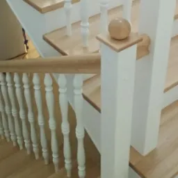 Jasne drewniane schody z białymi balustradami i drewnianą poręczą, zwieńczone kulą na słupku.