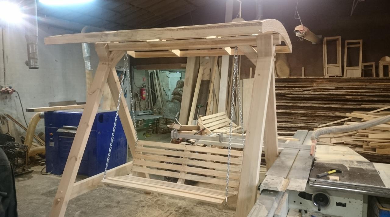 Drewniana huśtawka ogrodowa w trakcie montażu w warsztacie stolarskim, widoczne narzędzia i materiały budowlane w tle.