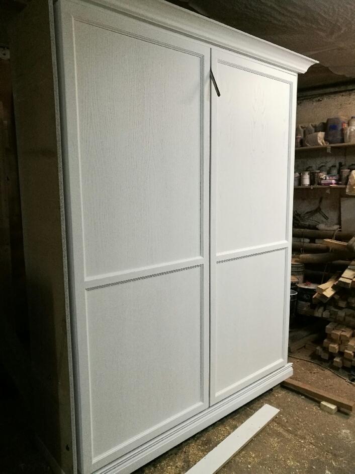 Biała, dwudrzwiowa szafa przesuwna z drewnianymi panelami, widoczna w warsztacie z narzędziami i materiałami w tle.