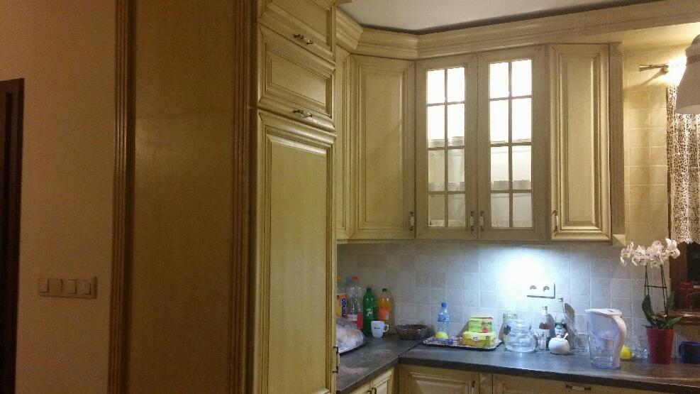 Klasyczne, kremowe szafki kuchenne z przeszklonymi górnymi szafkami, ciemny blat, białe płytki na ścianie i storczyk w doniczce.