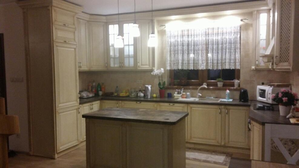 Jasna kuchnia w stylu rustykalnym z wyspą, okapem kominowym i oknem z firanką w kwiaty.