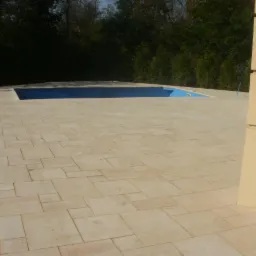 Jasne, nieregularne płyty wokół basenu, z widocznym fragmentem ściany budynku w tle. Ułożona kostka tworzy estetyczne otoczenie basenu.