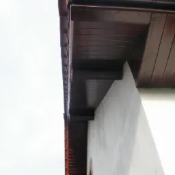 Detal elewacji budynku z brązowymi elementami drewnianymi i rynną. Widok z dołu, podkreślający połączenie drewna i jasnej ściany, z fragmentem okna w tle.