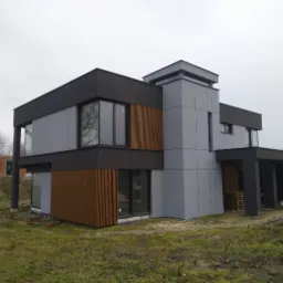 Nowoczesny dom jednorodzinny z elewacją w odcieniach szarości i brązu, widoczne elementy drewniane i duże okna, teren wokół w trakcie zagospodarowywania, w tle budynek w stanie surowym.