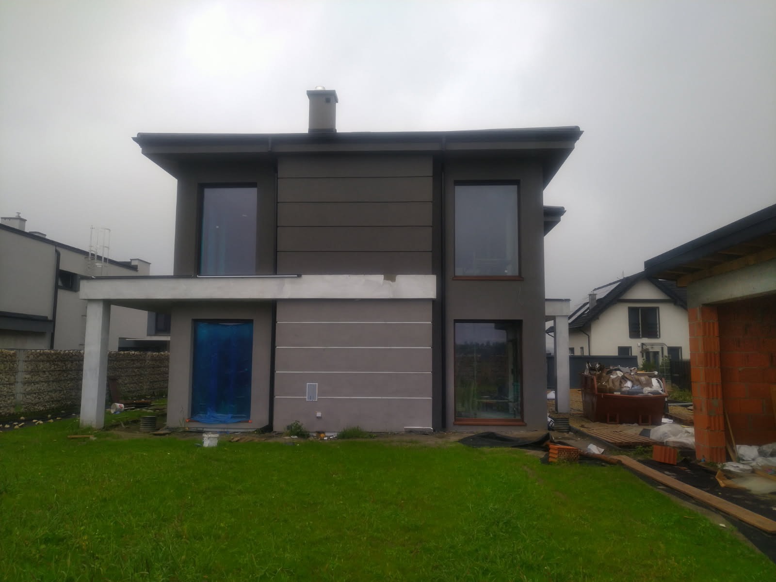 Nowoczesny dom dwukondygnacyjny z szarą elewacją w trakcie wykańczania, okna zabezpieczone folią, widoczny fragment trawnika i sąsiednie budynki w pochmurny dzień.