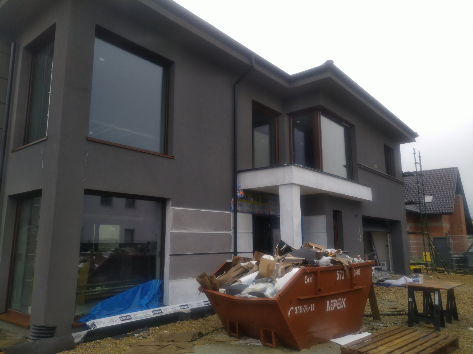 Dom jednorodzinny w trakcie wykończenia elewacji, widoczny kontener na gruz, zabezpieczona folią okienna stolarka i fragment ocieplenia pod balkonem.