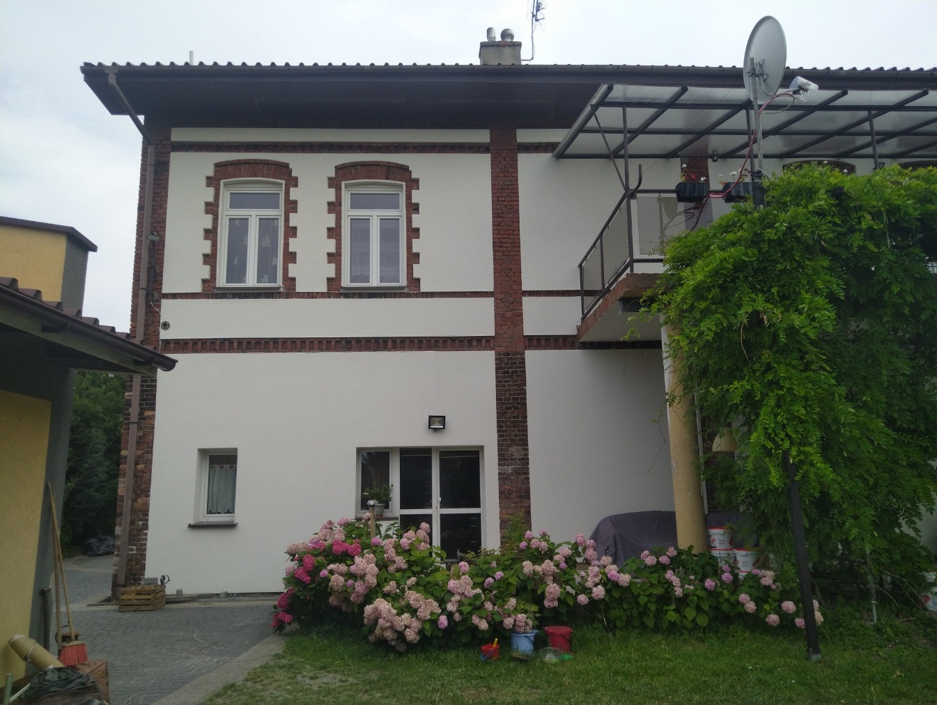 Ocieplony budynek z białymi ścianami i ceglanymi elementami dekoracyjnymi, widoczne kwiaty hortensji i balkon z zadaszeniem.