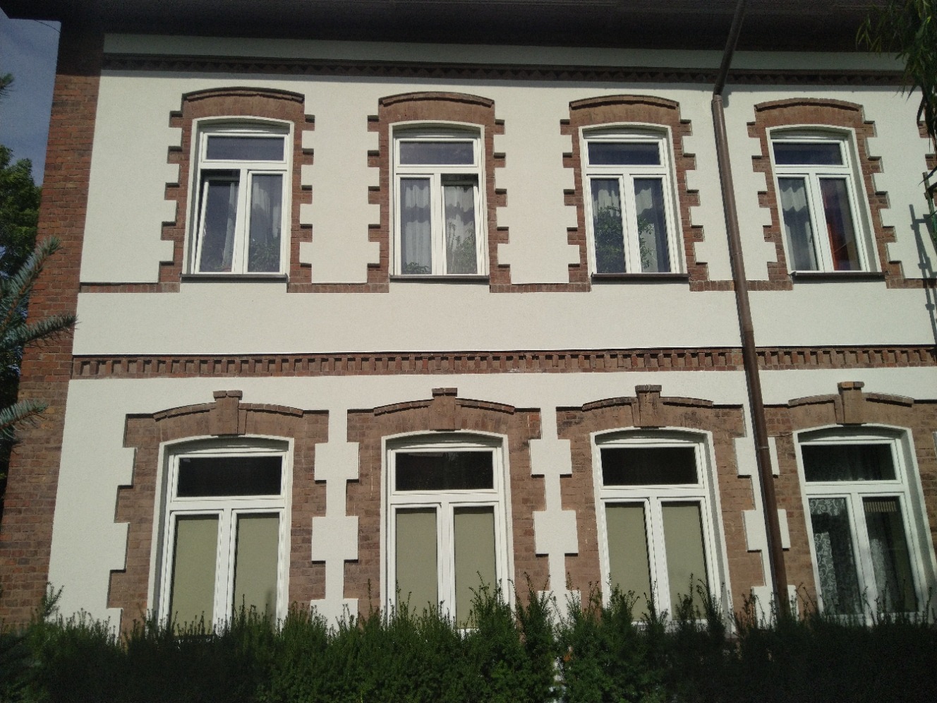 Odnowiona fasada budynku z białymi ścianami i ceglanymi detalami wokół okien, widok z poziomu gruntu na dwukondygnacyjny budynek.