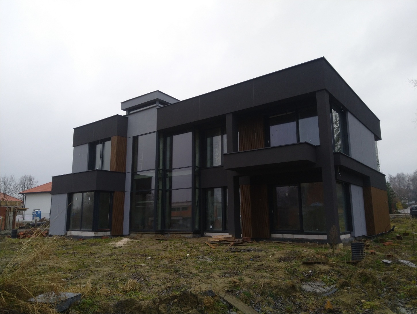Nowoczesny, dwukondygnacyjny dom z płaskim dachem w kolorach szarości i czerni, z dużymi oknami i drewnianymi akcentami, na działce w trakcie zagospodarowywania.