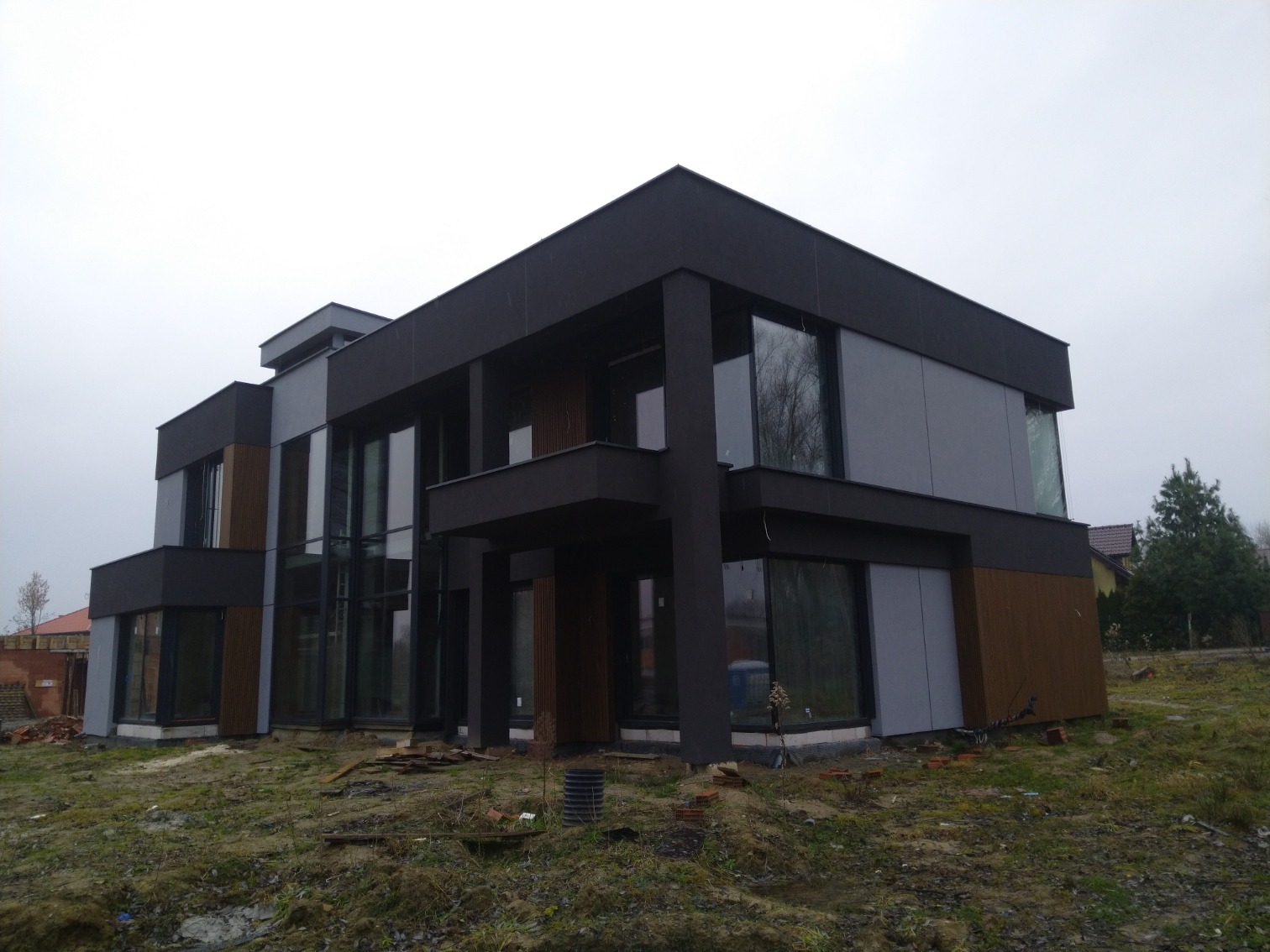 Nowoczesny, dwukondygnacyjny dom z płaskim dachem, elewacją w odcieniach szarości i czerni, z elementami drewnianymi, widoczny w pochmurny dzień na tle zieleni i sąsiedniej zabudowy, teren wokół...