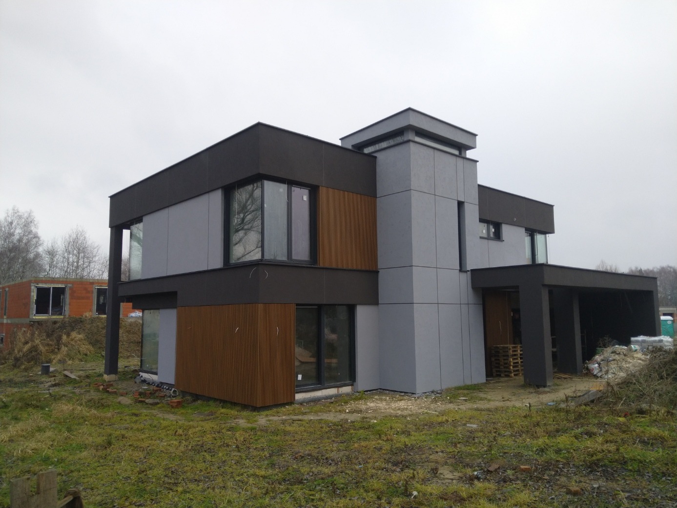 Nowoczesny dom jednorodzinny z elewacją w odcieniach szarości i brązu, widoczne elementy drewniane i duże okna, teren wokół w trakcie zagospodarowywania, w tle budynek w stanie surowym.