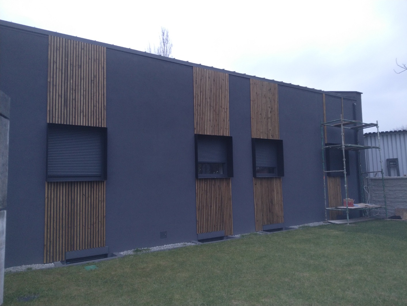 Elewacja budynku z szarego tynku i drewnianymi panelami pionowymi, okna z roletami zewnętrznymi, rusztowanie przy ścianie bocznej, trawnik przed budynkiem w pochmurny dzień.